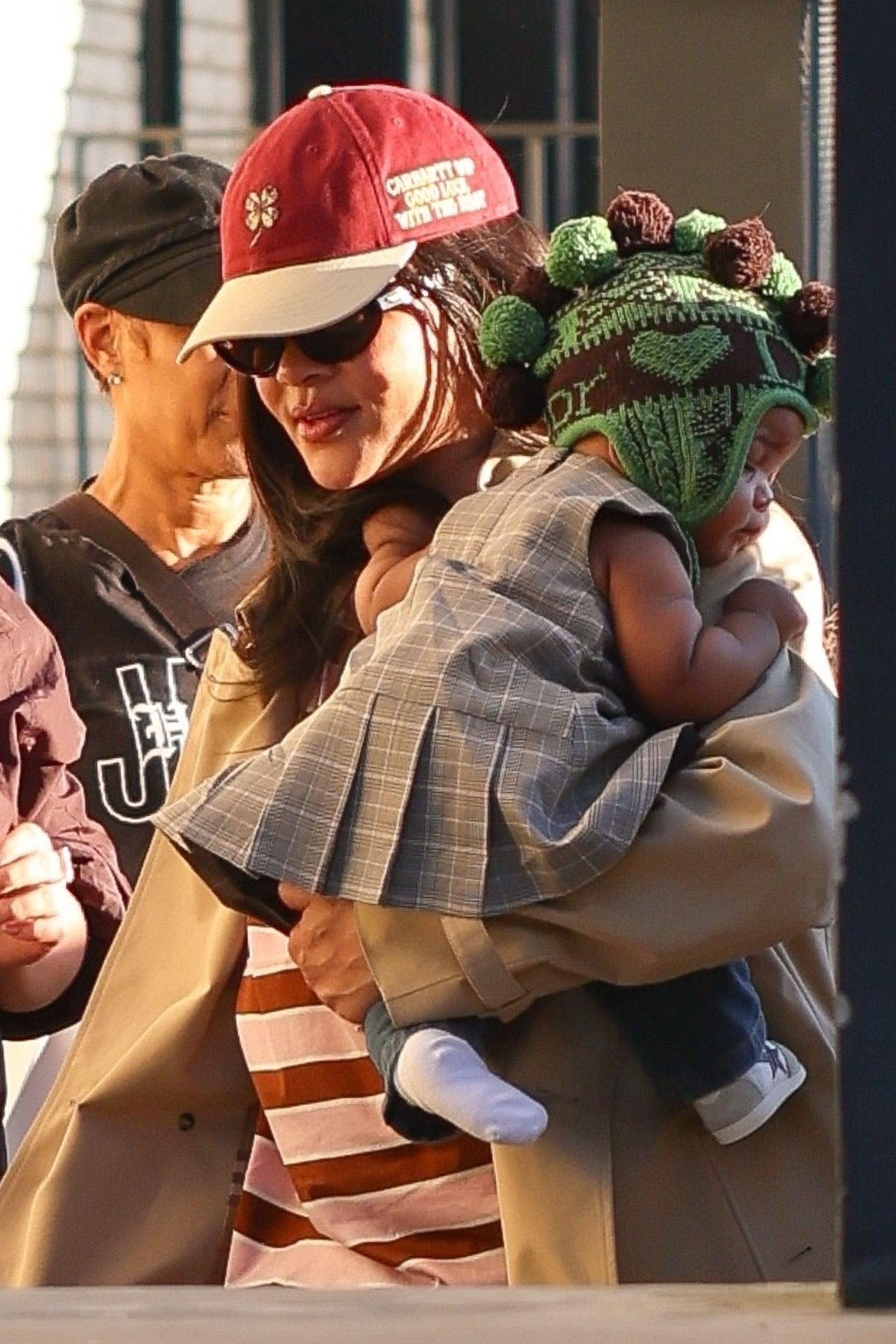 Rihanna wearing sunglasses, a red baseball cap and a tan trench coat, holding her baby who is wearing a plaid dress and a green knitted hat.
