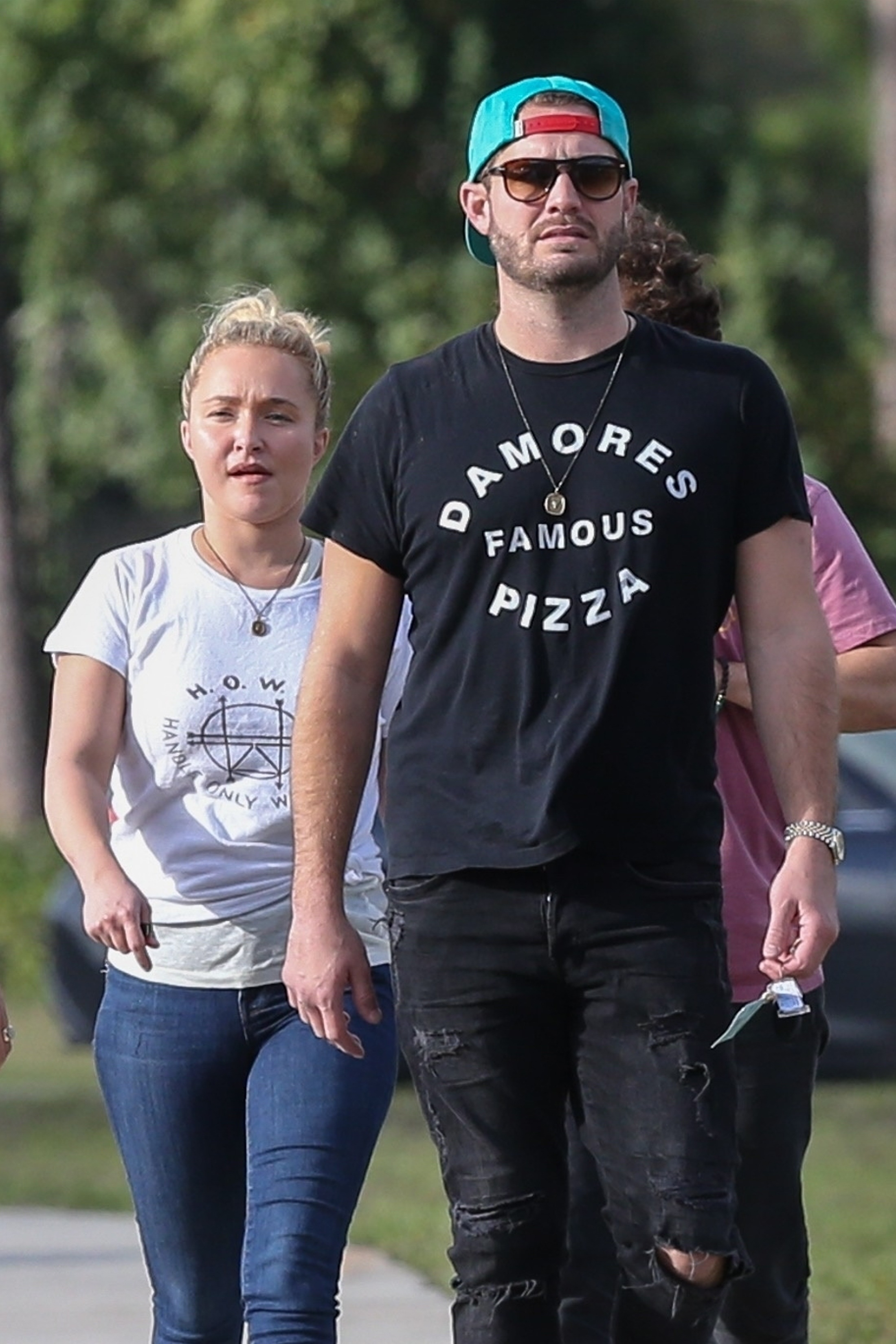 Hayden Panettiere and Brian Hickerson walk outdoors, with Panettiere wearing a white t-shirt and jeans, and Hickerson wearing a black t-shirt and ripped jeans with a backward teal cap and sunglasses.