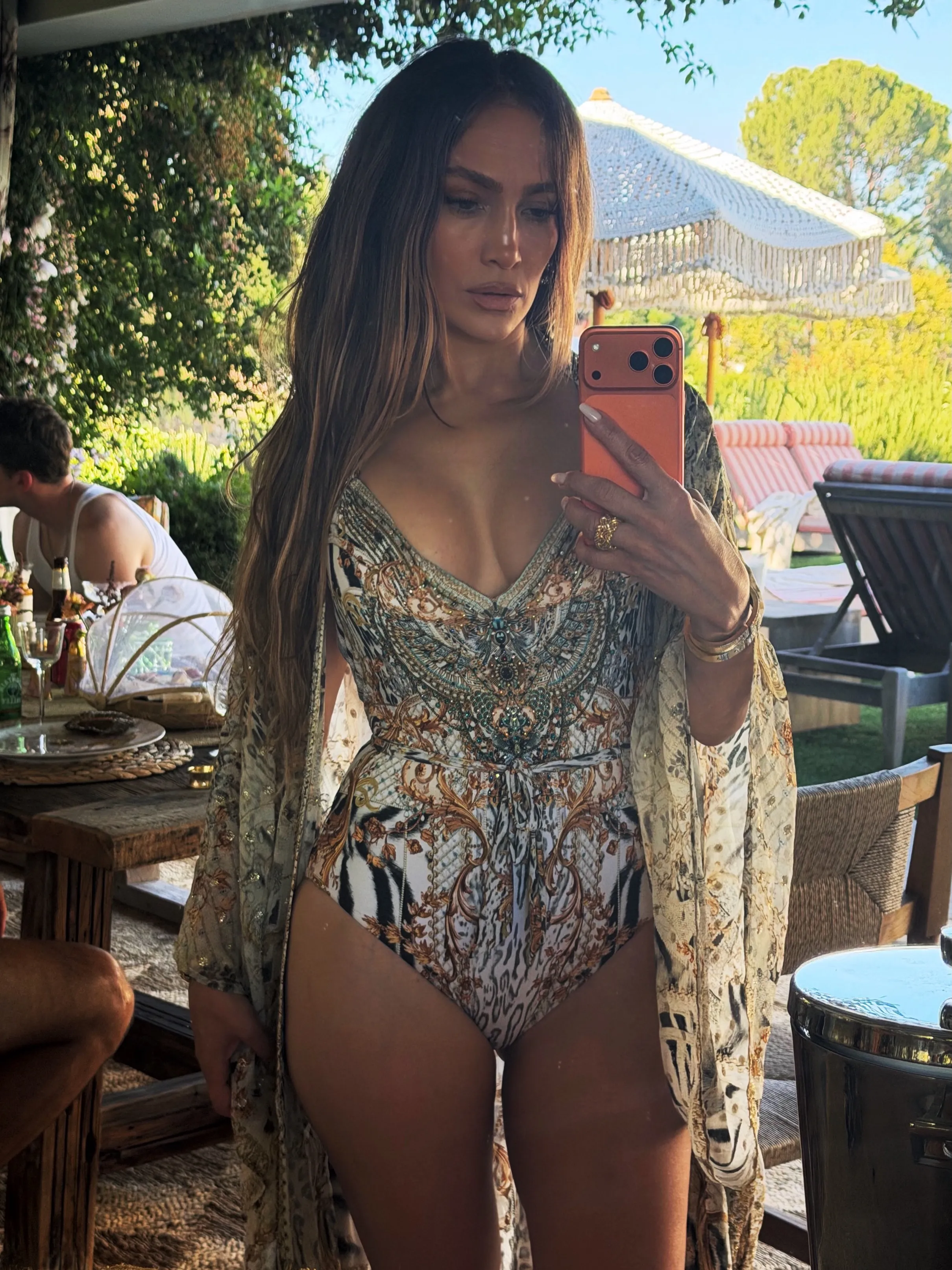 Jennifer Lopez taking a mirror selfie outdoors, wearing a patterned swimsuit and a sheer cover-up.