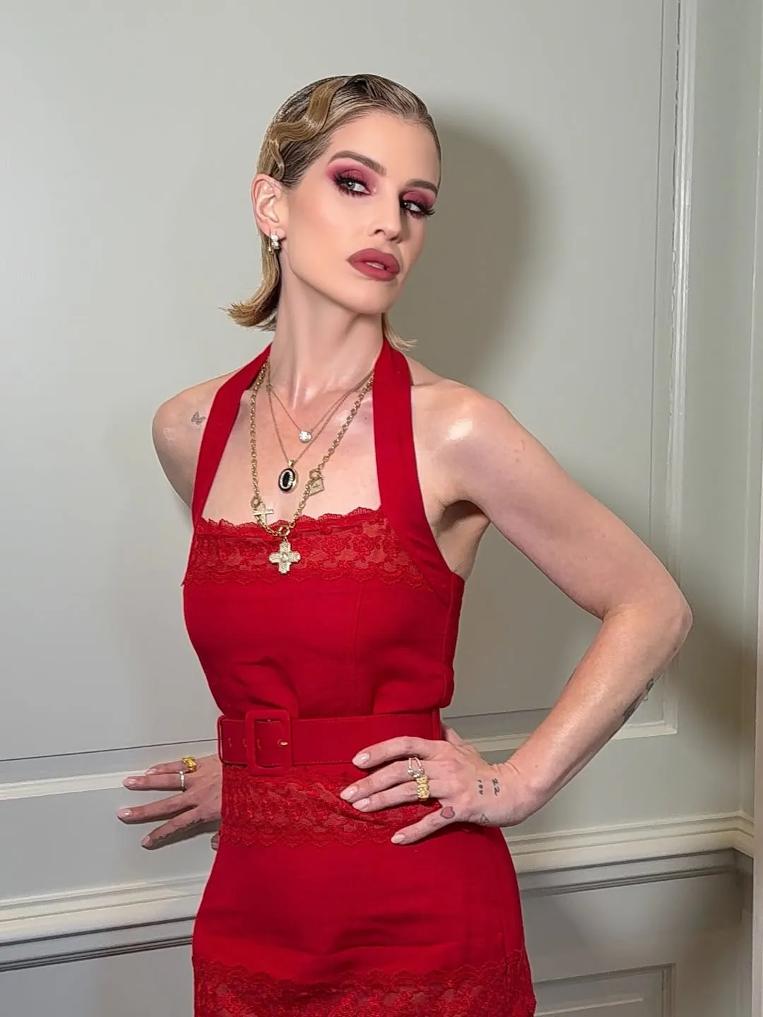 Kelly Osbourne in a red halter dress with a belted waist and lace trim, featuring deep pink eyeshadow and wavy blonde hair, posing with hands on hips.