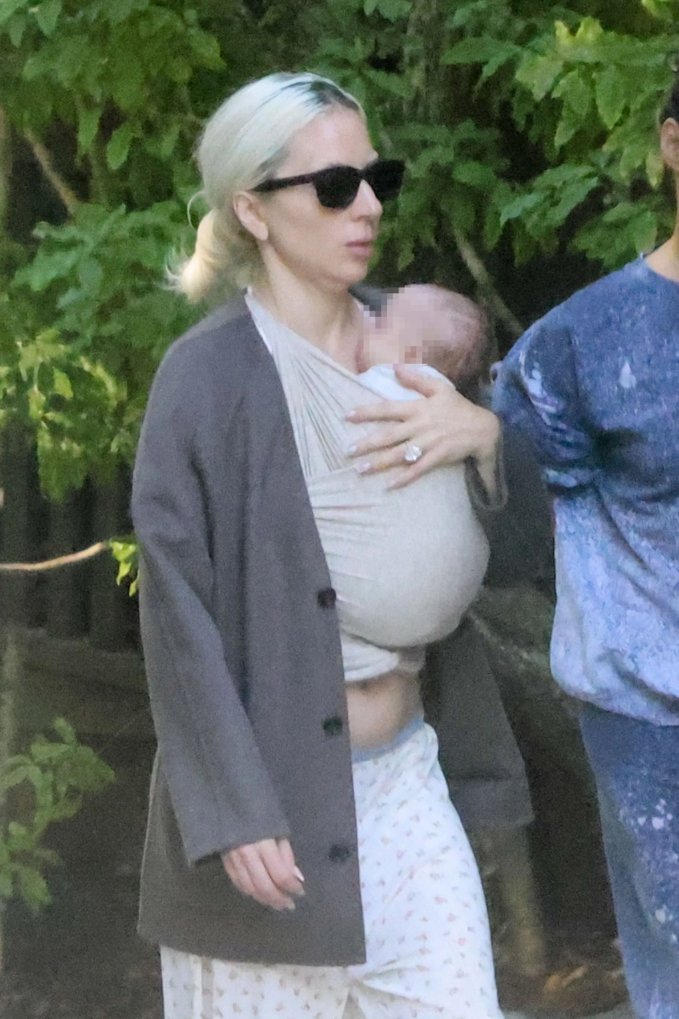 Lady Gaga wearing sunglasses with a baby in a carrier and a diamond ring on her left hand.