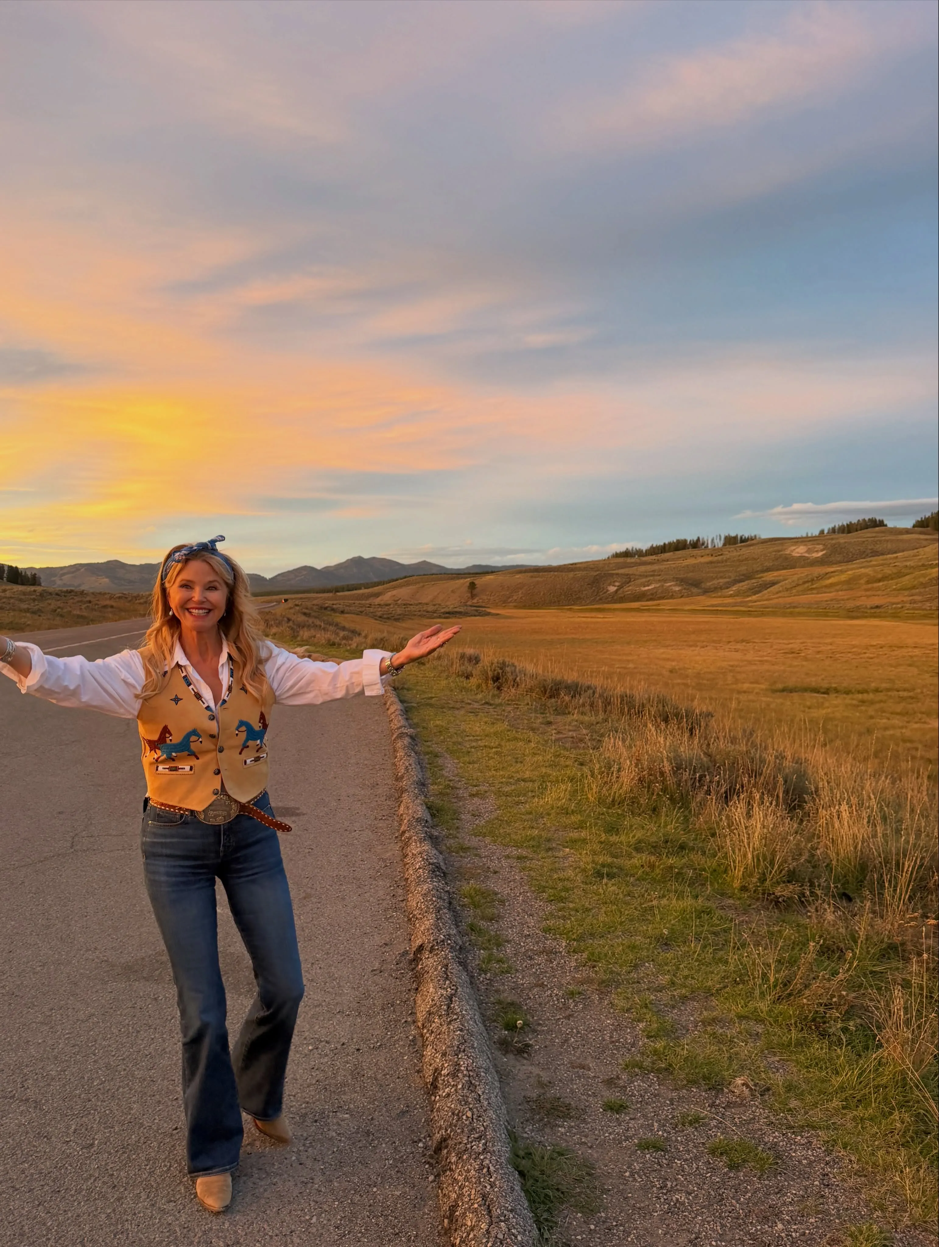 Christie Brinkley smiling with arms outstretched in a cowboy vest and jeans on a road in a field at sunset.