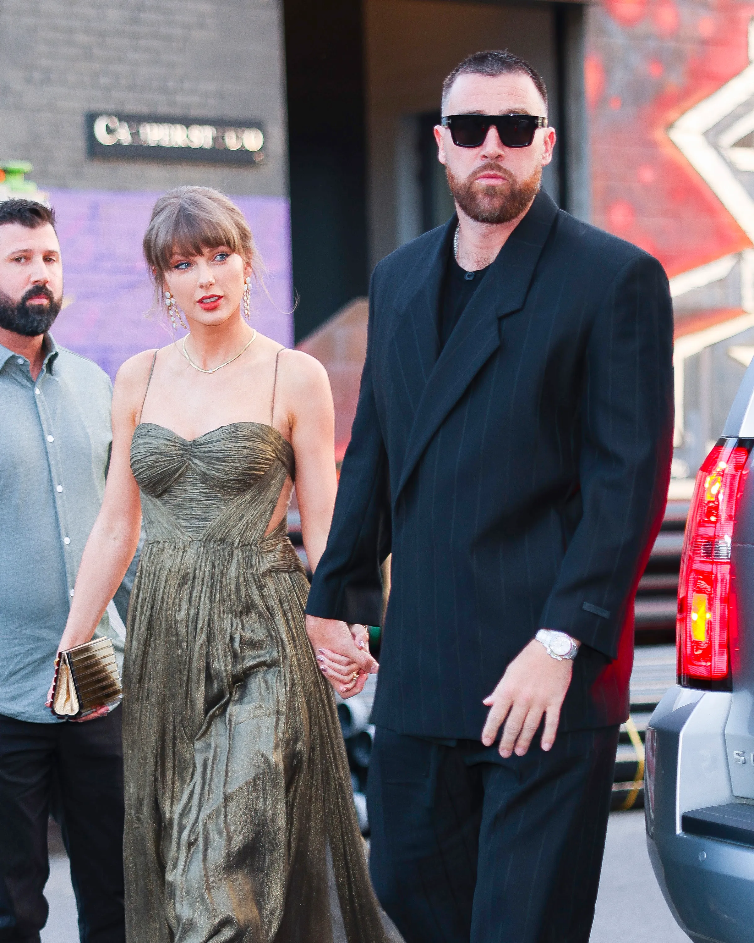 Taylor Swift in a gold dress holding hands with Travis Kelce in a dark pinstripe suit and sunglasses.