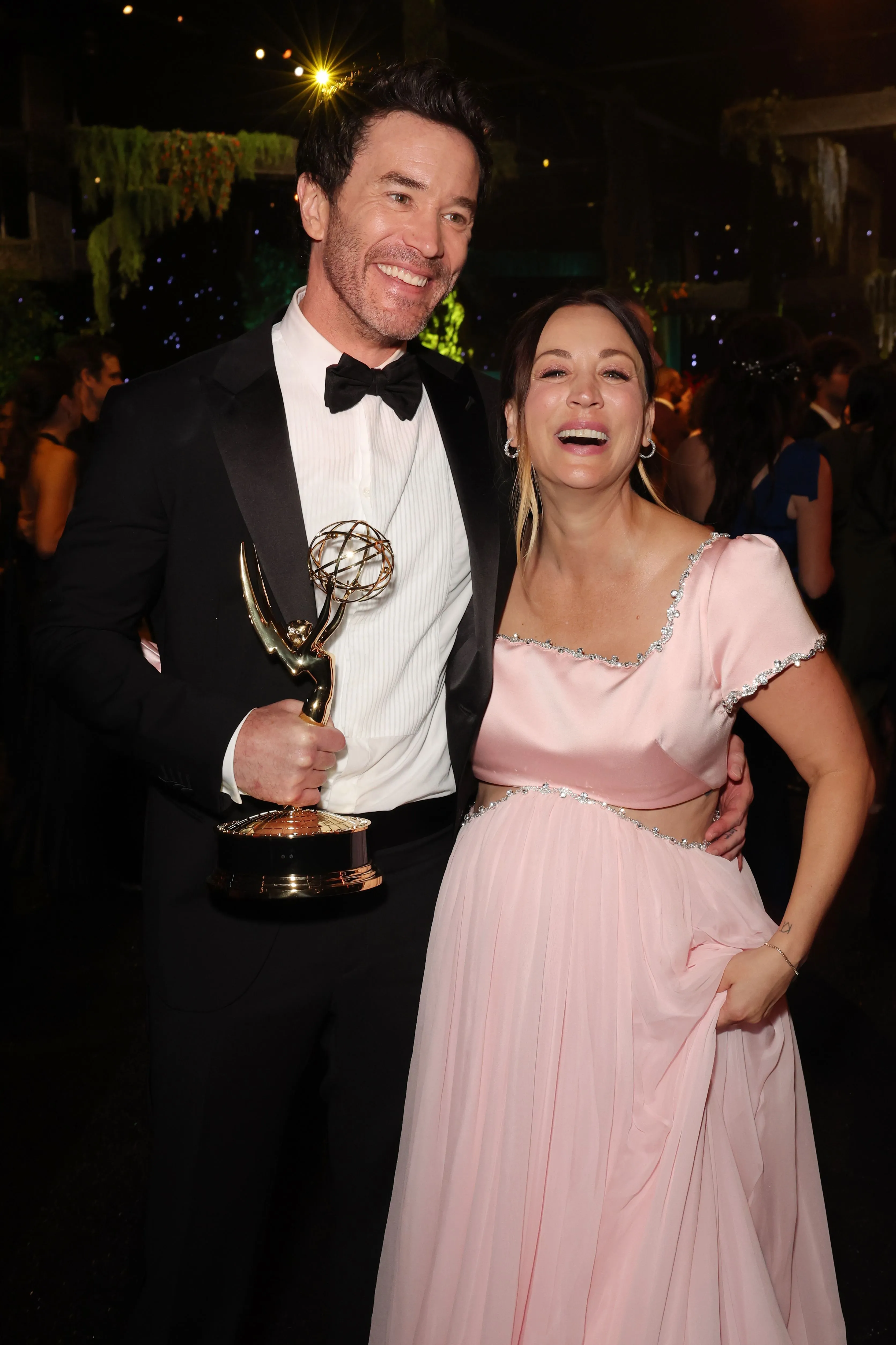 Tom Pelphrey in a tuxedo holding an Emmy award, with Kaley Cuoco in a pink dress, laughing beside him.