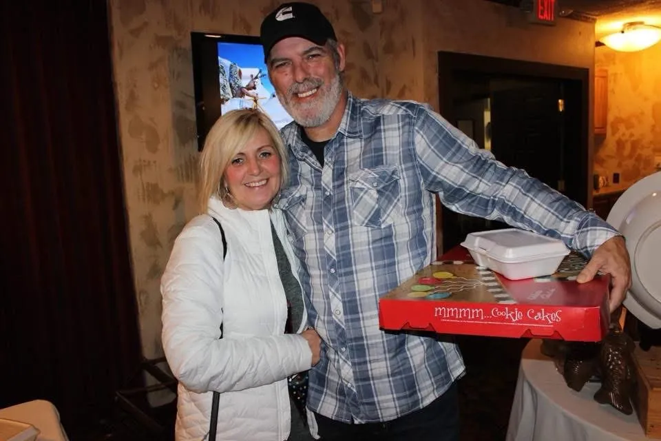 A smiling woman with blonde hair stands next to a smiling man with a beard and a black hat who is holding a "mmmm.Cookie Cakes" box.