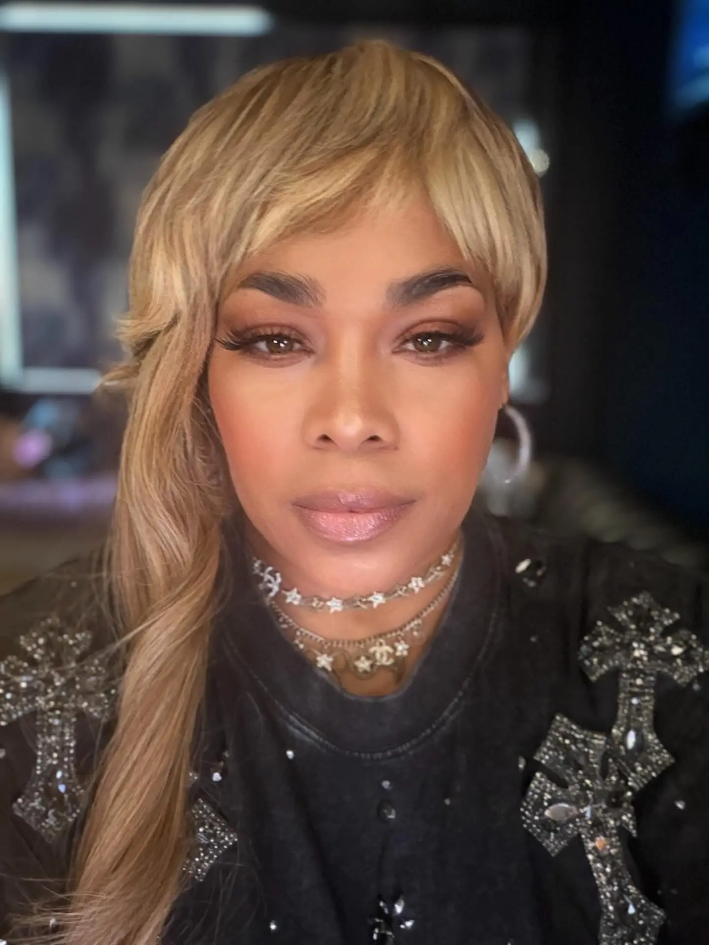 T-Boz Watkins looking at the camera, wearing a blonde wig, layered necklaces, and a black top with embellished crosses.