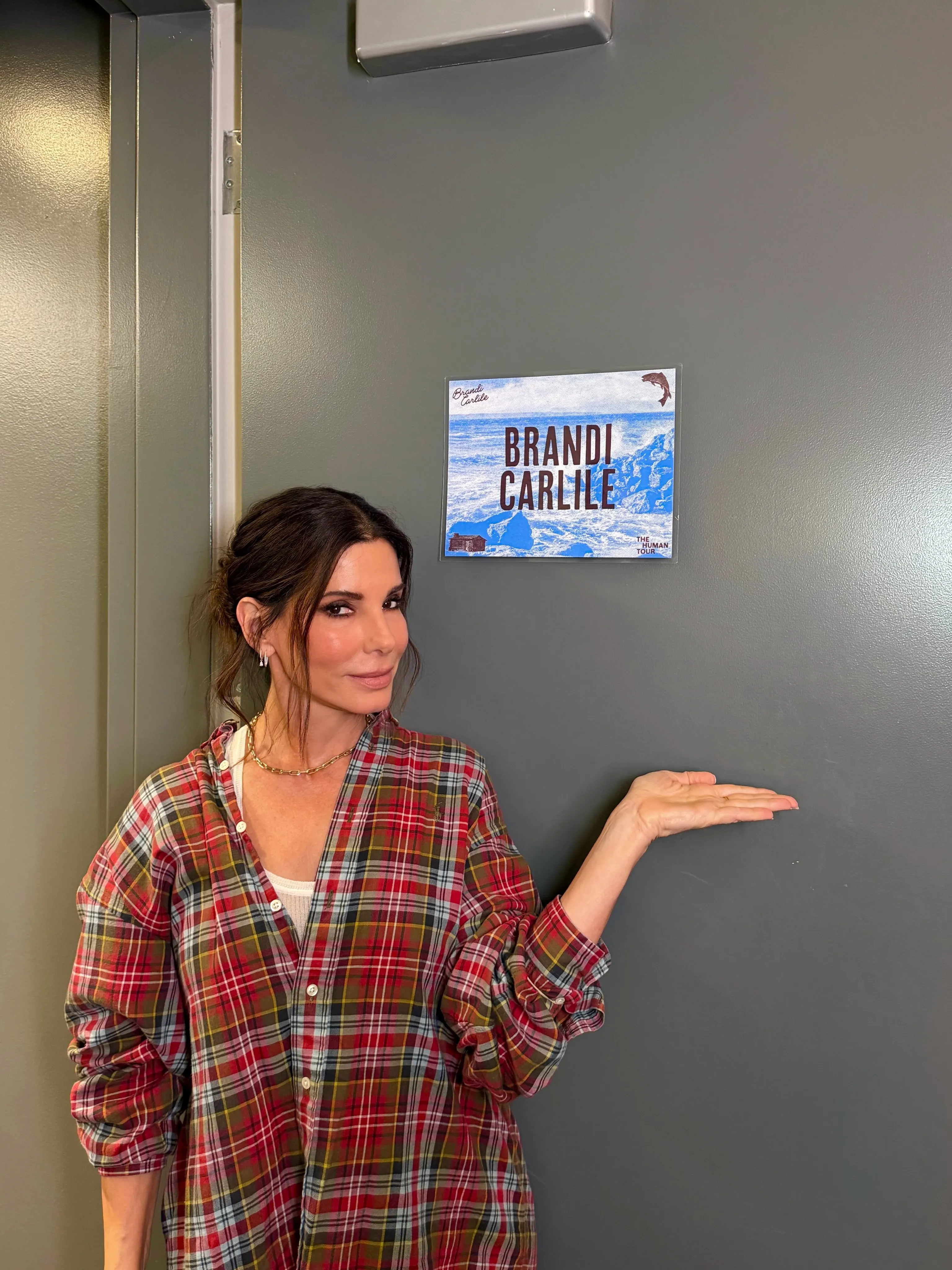 Sandra Bullock in a red and green plaid shirt gestures to a "Brandi Carlile The Human Tour" poster on a gray wall.