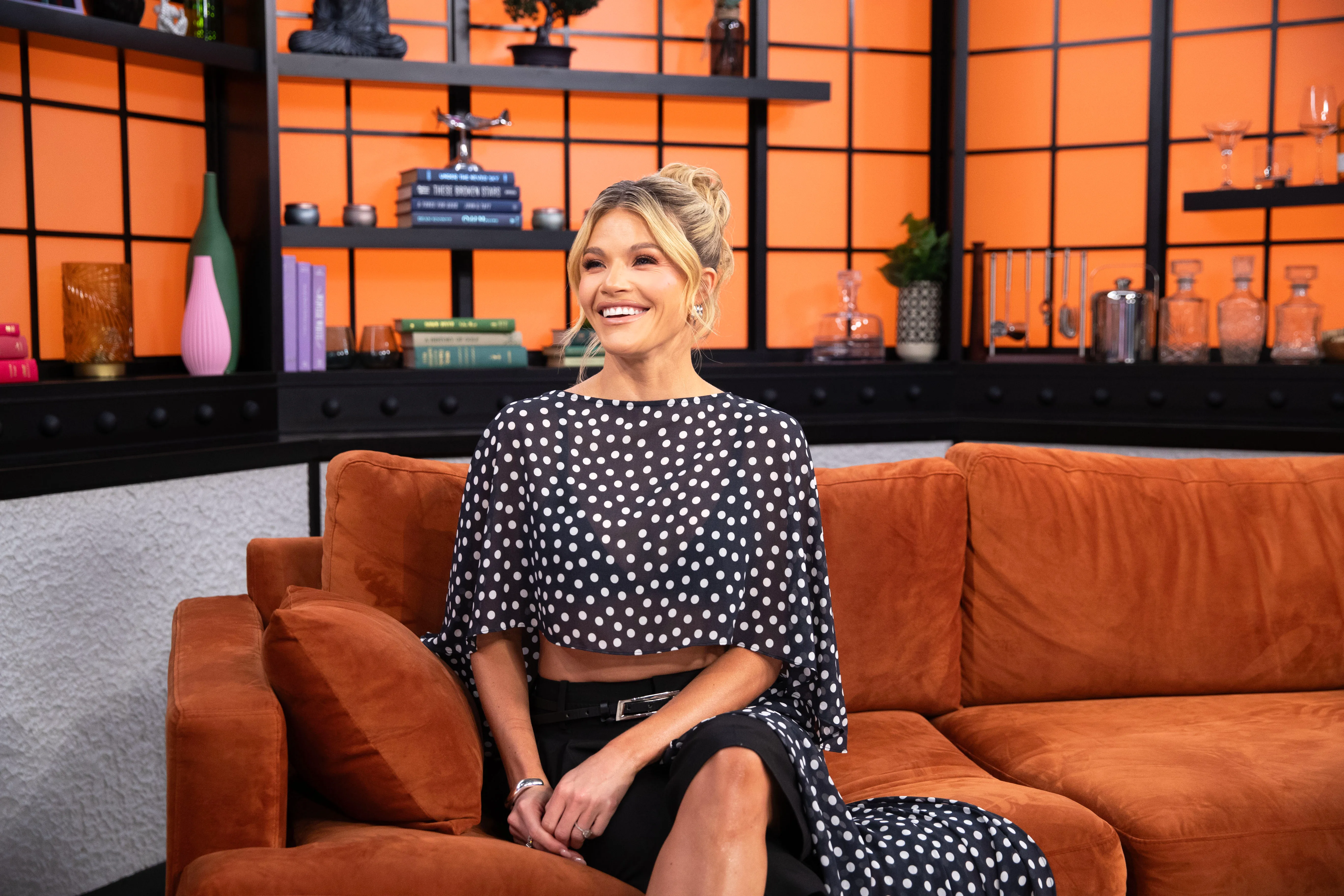 Witney Carson sitting on an orange couch in a studio, wearing a black and white polka dot top and skirt.
