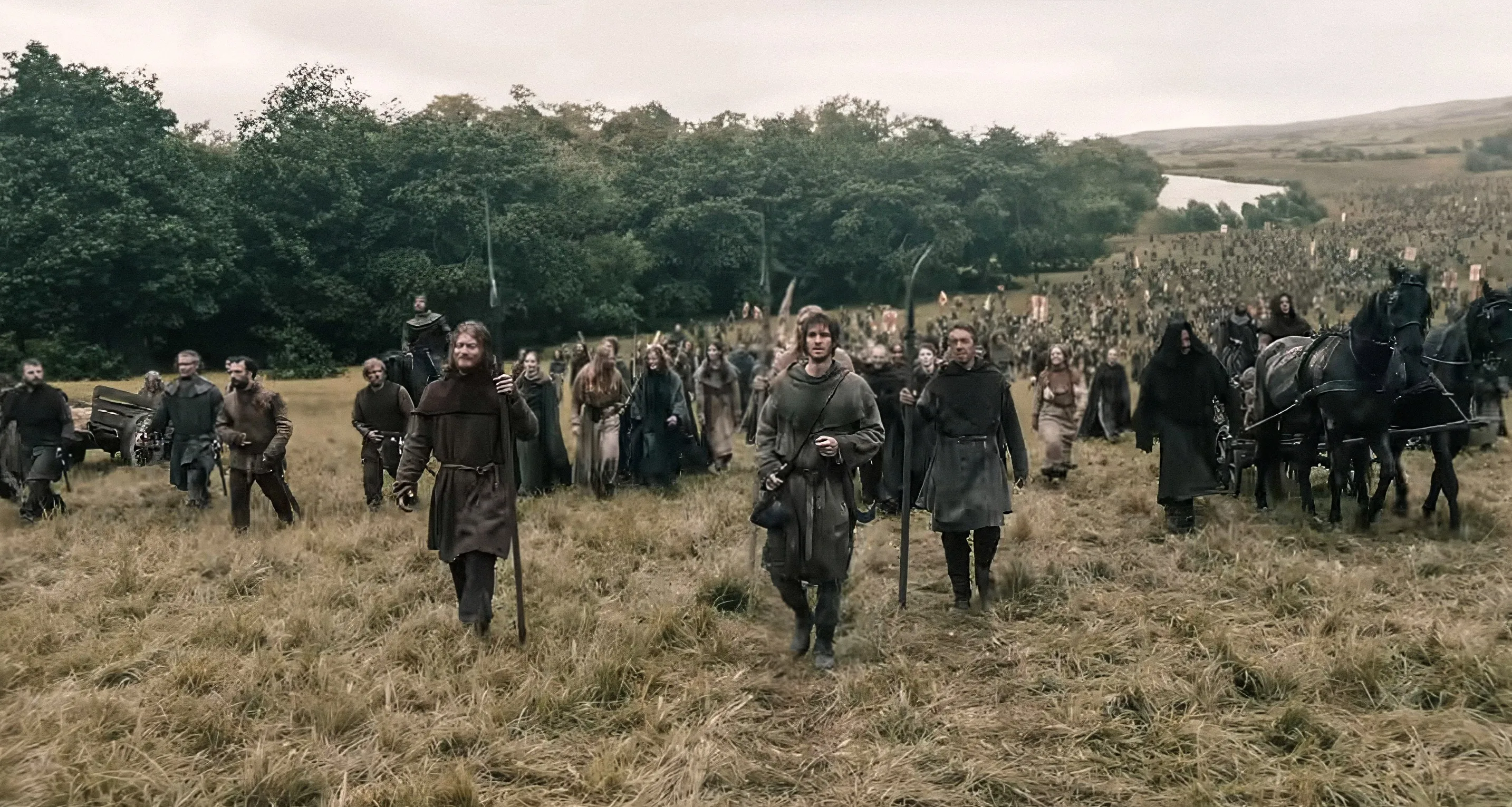 Andrew Garfield, in medieval attire, leading a large group of people across a grassy field.