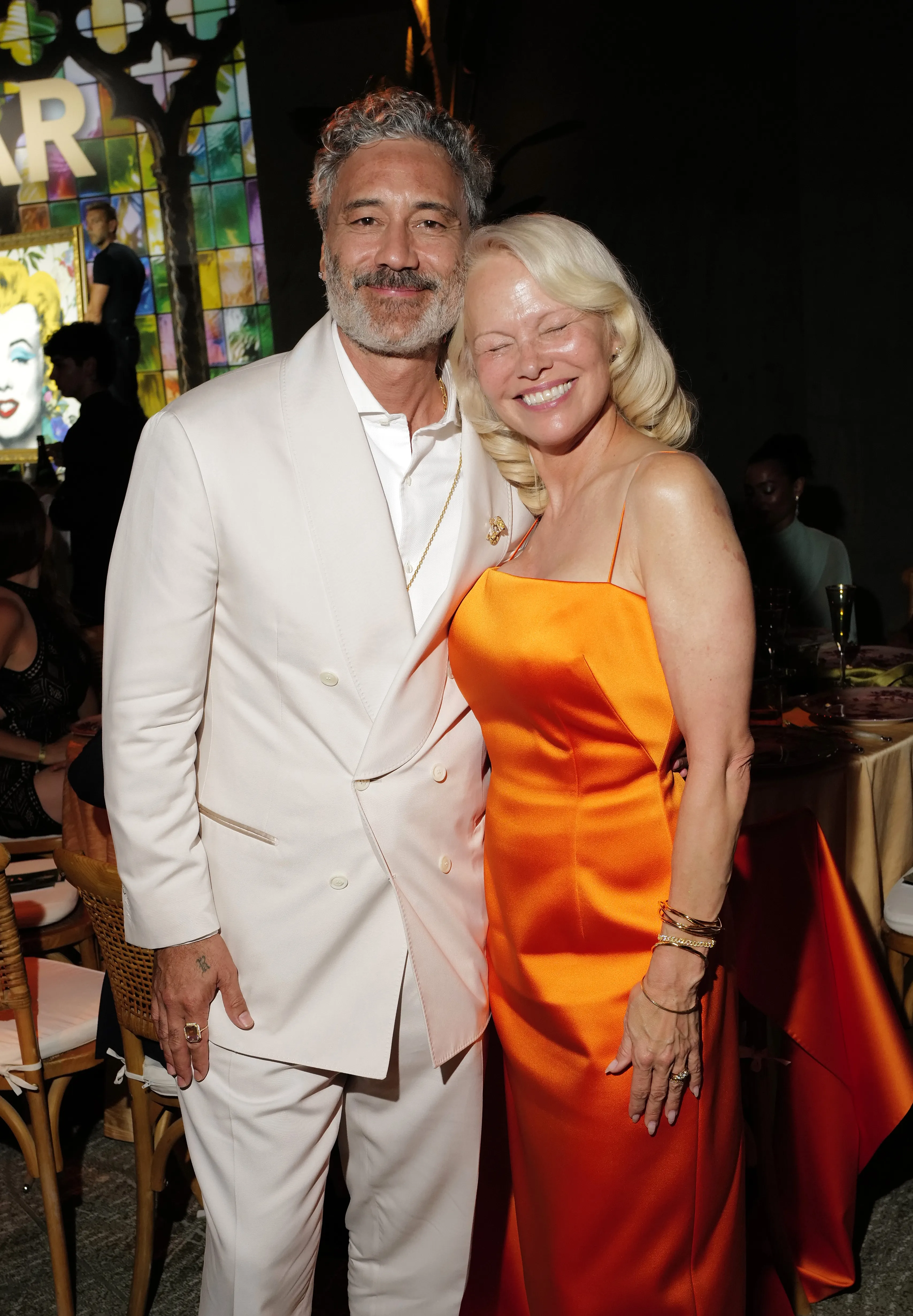 Taika Waititi in a white suit and Pamela Anderson in an orange dress posing together.
