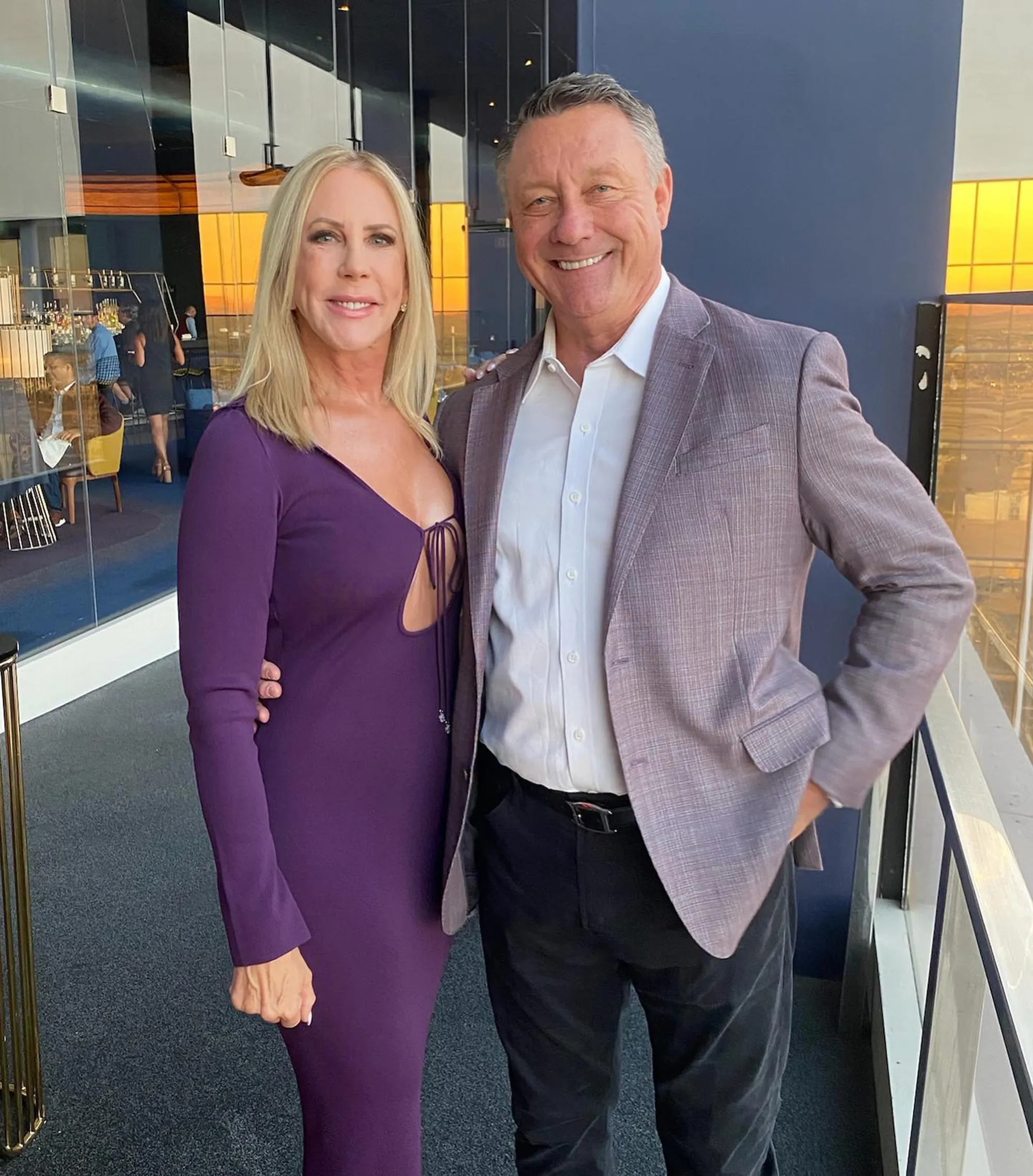 A smiling man in a suit jacket and a smiling woman in a purple dress standing arm in arm.